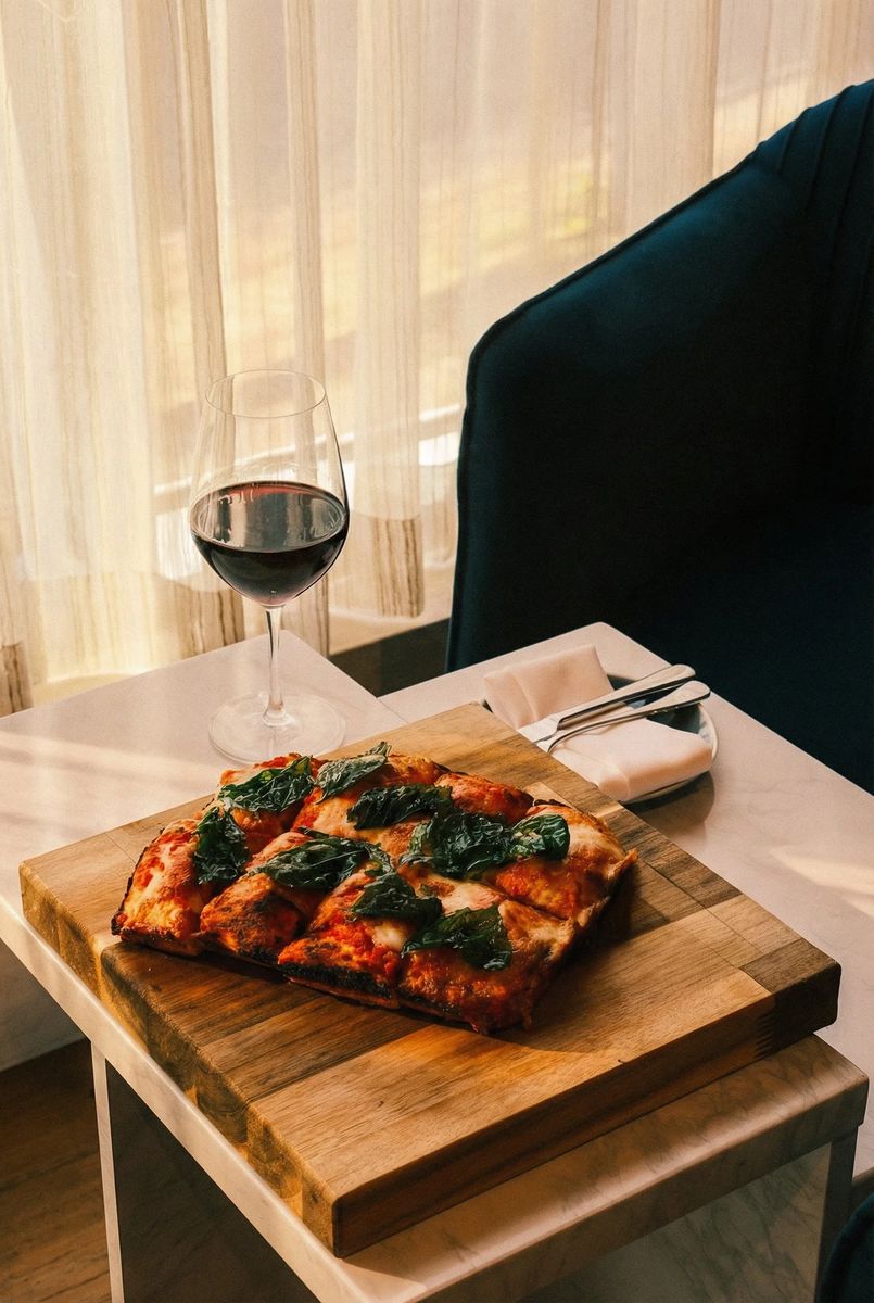 Roman-style al taglio pizza topped with basil on a wooden board beside a glass of red wine at Provisions Italiana
